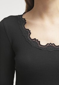 Musta ribbed pitkähiha kauluspaita, jossa on vaaleat reunukset ja tummat yksityiskohdat, korostaen tekstuuria ja rakenteellista muotoa.