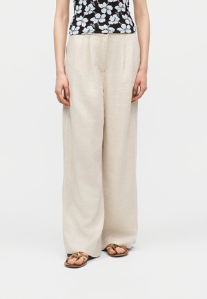 Pantaloni largi bej din in purtați cu sandale maro cu bretele încrucișate și un top fără mâneci negru cu imprimeu floral.