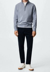 Homme portant un pull à demi-zip gris clair, un t-shirt blanc, un pantalon noir et des baskets blanches, se tenant contre un fond neutre.