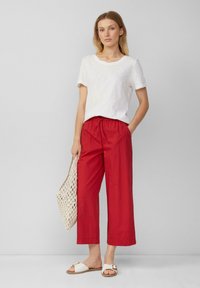Pantalons larges rouges avec taille élastique, associés à un haut court à manches courtes de couleur claire et texturé, et un sac en osier. Sandales à glissière blanches.