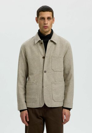 Homme portant une veste beige à carreaux par-dessus un col roulé noir et un pantalon marron, debout devant un fond clair uni.