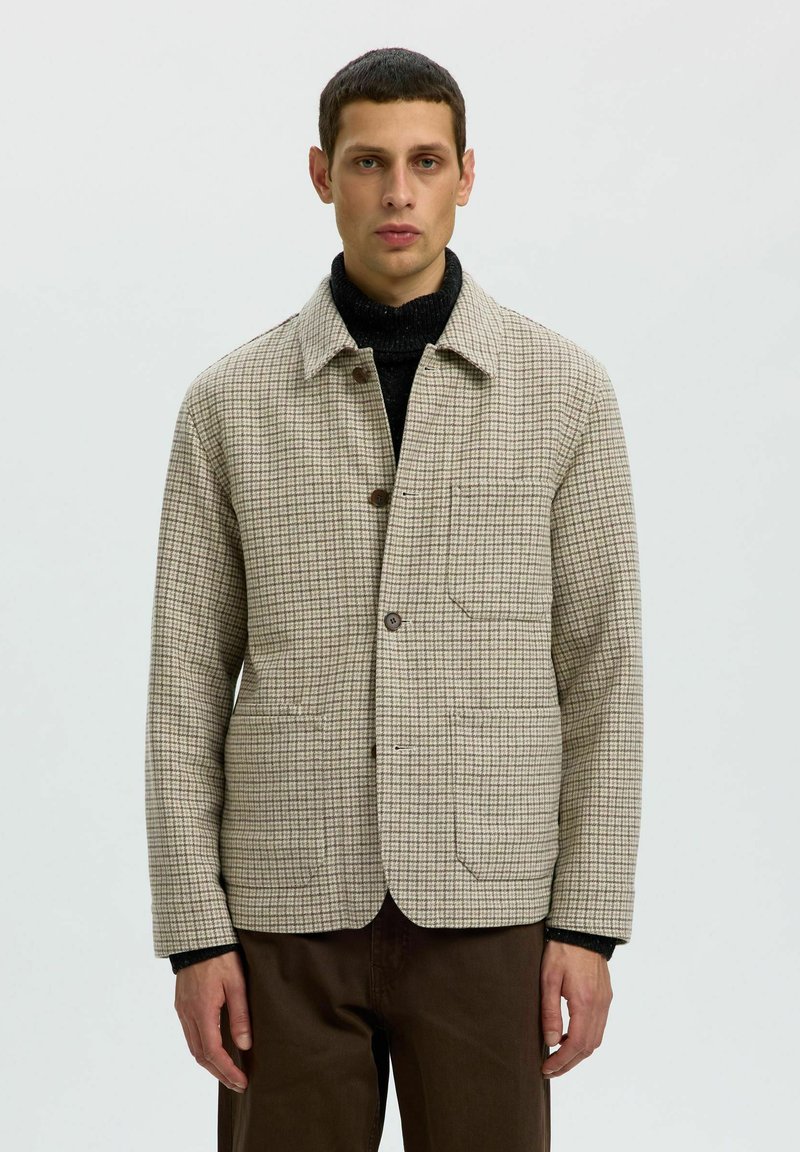 Homme portant une veste beige à carreaux par-dessus un col roulé noir et un pantalon marron, debout devant un fond clair uni.