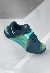 Nike sportssko i teal, navy og gulgrøn mesh med snørebånd, logo og struktureret sål på en lys grå overflade.