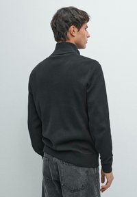 Maglione pullover nero in un tessuto morbido e lavorato a maglia. Presenta un collo alto e polsini a coste, indossato con jeans in denim grigio.
