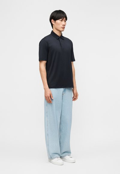 Άνδρας όρθιος, με μαύρο πόλο πουκάμισο, ανοιχτό μπλε παντελόνι με φαρδιά μπατζάκια και λευκά αθλητικά παπούτσια, μπροστά σε απλό ανοιχτό φόντο.