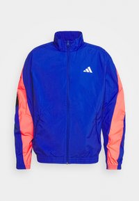 Chaqueta ligera azul con mangas rosas y negras, cuello alto, cierre frontal con cremallera y un logo blanco bordado en el pecho.