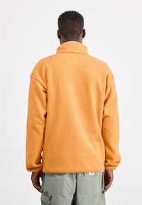 Orange fleecepullover med hög krage, texturerad yta, sänkt axelsömmar och bakloopdetalj. Bärs med cargobyxor.