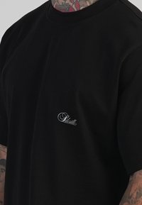 Černé bavlněné tričko s volnějším střihem, s malým bílým logem na hrudi a klasickým kulatým výstřihem. Bez výrazných vzorů nebo zdobení.