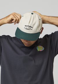 Bílá čepice se zeleným kšiltem, s černým textem a logem na přední straně. Nositel upravuje čepici a má na sobě tmavé tričko.