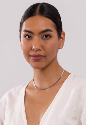 Silberne Metallkette mit geschwungenem Design, verfügt über eine glatte, polierte Oberfläche. Getragen um den Hals eines Models, das eine weiße Bluse trägt.