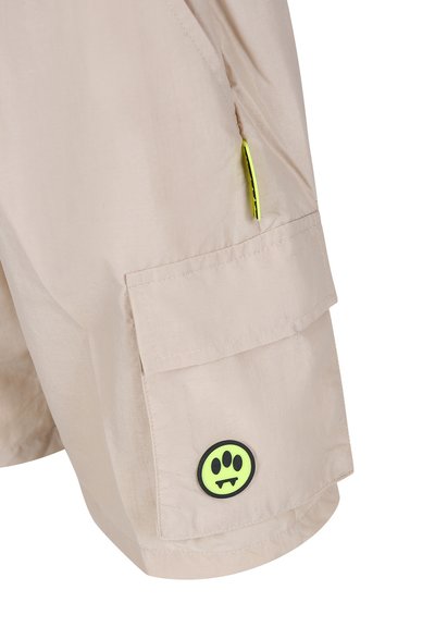 Pantaloni cargo beige con una texture liscia, dotati di una tasca laterale e di un distintivo circolare verde con un design nero. Accenti neon notevoli.