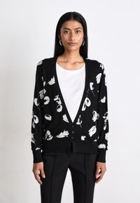 Svart cardigan med vit blommönster, V-ringning, knäppning fram, lång ärm och avslappnad passform, ihop med en vit topp och svarta byxor.