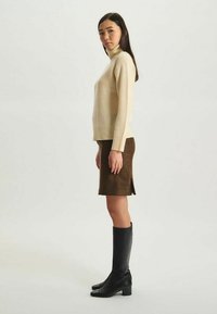 Pull à col roulé beige, au design ajusté. Jupe brune à motifs avec une fente sur le côté. Bottes noires jusqu'aux genoux avec un petit talon.