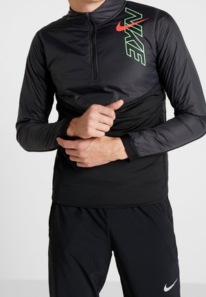 Mann in schwarzem Nike-Pullover mit neongrünem und rotem Logo, Hände verschränkt auf der Taille, trägt schwarze Sporthose mit weißem Nike-Swoosh.