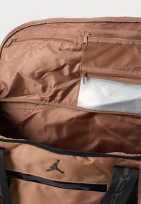 Interior de uma bolsa de tecido castanho com textura lisa, apresentando compartimentos com fecho éclair e um bolso em malha. Logótipo preto no exterior.