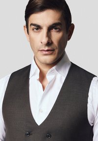 Ciemnoszary kamizelka z dekoltem w serek na białej koszuli z kołnierzykiem, z dwoma czarnymi guzikami i gładką fakturą. Neutralne tło.