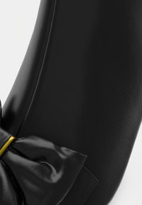 Gros plan d’une chaussure en cuir noir avec un grand nœud en cuir et un petit accent doré près du centre du nœud.