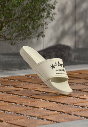 Șlapi bej Karl Lagerfeld cu logo scris negru pe o suprafață de gresie din lemn și o plantă verde neclară în fundal.