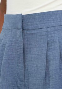 Μπλε παντελόνι ψηλόμεσο, ραμμένο με φερμουάρ μπροστά κρυφό και πιέτες, φορεμένο με λευκό τοπ που είναι μαζεμένο μέσα στο παντελόνι.