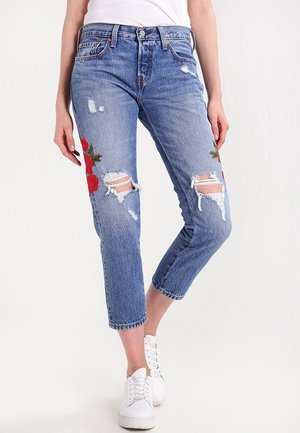 Femme portant un jean bleu taille mi-haute déchiré avec une broderie florale rouge sur les cuisses et des baskets blanches, debout devant un fond uni.