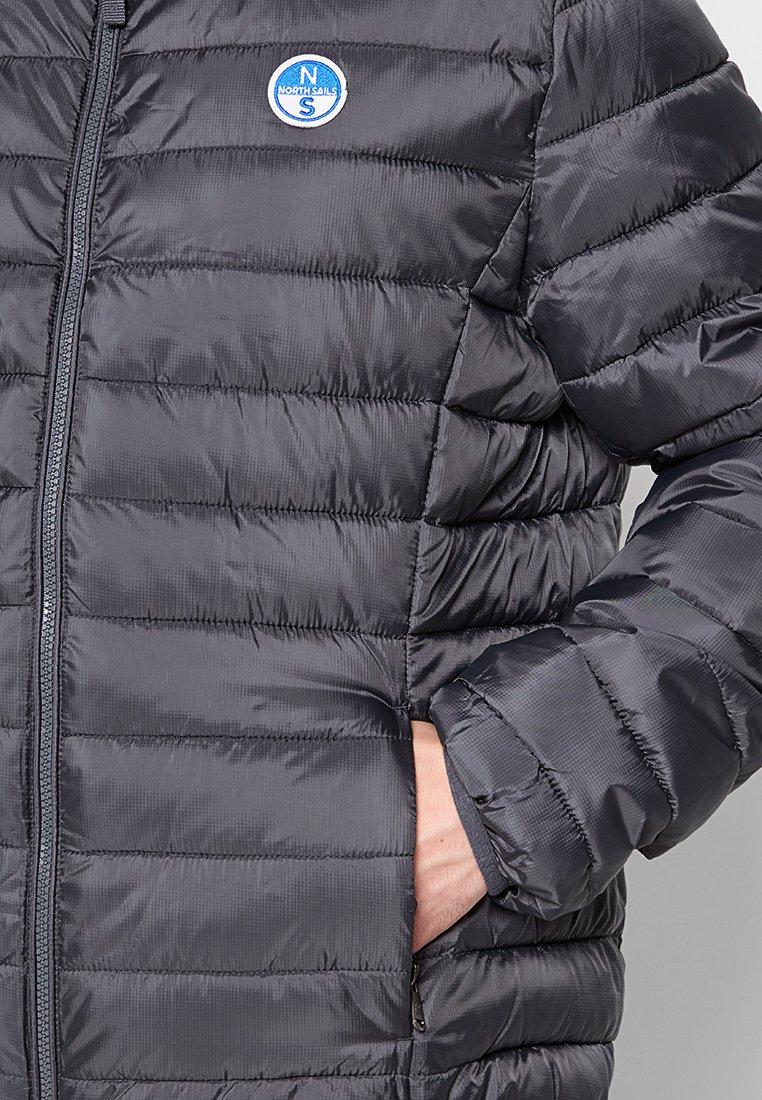 Giacca a vento grigia scura con texture trapuntata, chiusura con zip e tasche laterali. Presenta un logo blu tondo sulla parte superiore sinistra.