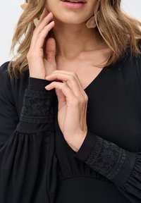 Kobieta w czarnej bluzce z długim rękawem i koronkowymi kwiatowymi mankietami, dotykająca swojego nadgarstka, z widoczną podbródkiem, ustami i falującymi, jasnobrązowymi włosami.