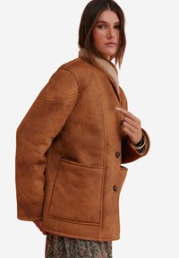 Vrouw in een bruine schapenvachtjas met knopen over een bloemenrok, die naar het borstgedeelte van de jas wijst.