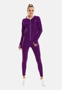 Junge Frau steht selbstbewusst, trägt einen lila Kapuzenpullover mit Reißverschluss und passende Leggings sowie weiße Turnschuhe vor einem schlichten Hintergrund.