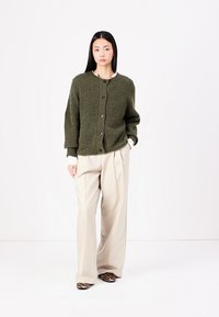 Cardigan tricotat texturat, de culoare verde măsliniu, cu guler rotund, închidere cu nasturi și croială supradimensionată, asociat cu pantaloni largi bej și pantofi cu imprimeu animalier.