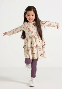 Bloemenjurk met een gelaagd ontwerp in crème, met paarse en groene accenten. Gecombineerd met geribbelde paarse legging en witte schoenen.