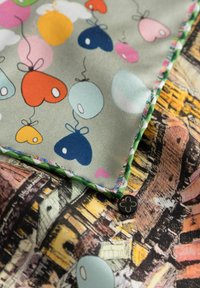 Kleurrijke stof met harten en ballonnen op een lichte achtergrond, met een groene patroonrand en een zwarte decoratieve knop in de hoek.