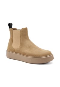 Botín Chelsea de ante marrón con paneles laterales elásticos y una lengüeta negra. Presenta una suela de goma gruesa de color beige con detalles texturizados.