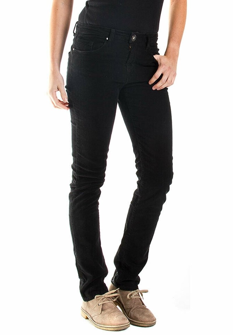 Personne portant un jean noir slim et des chaussures à lacets en daim beige, debout avec une main posée sur la hanche.
