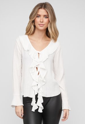 Mujer joven con blusa blanca de manga larga y lazos fruncidos en la parte delantera, y pantalones de cuero negro, posando frente a un fondo liso.