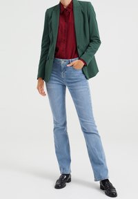 Mörkgrön kavaj över en röd knapparskjorta, kombinerad med ljusblå jeans och svarta loafers; strömlinjeformad silhuett, mjuka texturer.