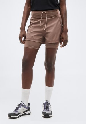 Persoană purtând pantaloni scurți maro în straturi cu șnur, șosete albe până la gleznă și pantofi gri pentru alergare pe trasee, stând în fața unui fundal simplu și deschis la culoare.