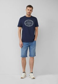 Man staat tegen een grijze muur, gekleed in een marineblauw graphic T-shirt, lichtblauwe denim shorts en witte sneakers met beige zolen.