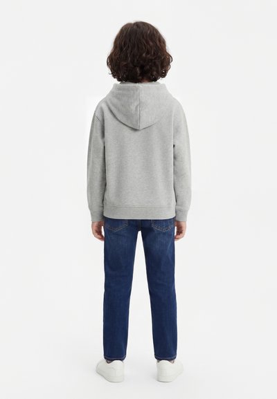 Child standing facing away wearing a gray hoodie, blue jeans, and white sneakers against a plain white background.