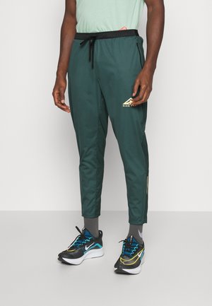 Homme portant un pantalon de survêtement Nike vert foncé avec une ceinture noire et des chaussures de course Nike noires, bleues et jaunes sur un fond uni.