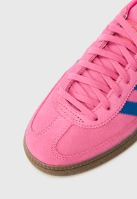 Ροζ σουέτ αθλητικό παπούτσι με μπλε λεπτομέρειες, διαθέτει υφασμένη ελαστική σόλα, επίπεδα ροζ κορδόνια και ραφές κατά μήκος του μπροστινού μέρους και των πλευρών.