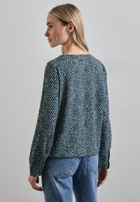 Maglietta a maniche lunghe, in tessuto texture, di colore verde scuro con un motivo astratto bianco e azzurro chiaro; scollatura rotonda; abbinata a pantaloni in denim blu.
