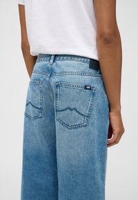 Gaiši zili, plaši džinsi ar izbalinātu apdari. Iezīmē divas aizmugures kabatas ar kalna formas šuvju detaļu un melnu jostas tagu.