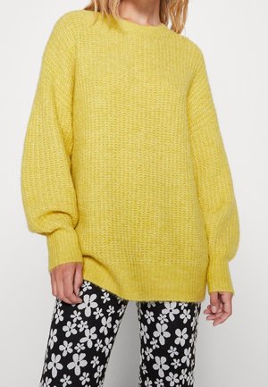 Femme portant un pull en maille jaune ample et un pantalon noir avec un motif floral blanc sur un fond uni.