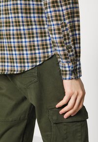 Camisa de franela con un diseño a cuadros en verde, azul y naranja, combinada con pantalones cargo verde oliva que tienen bolsillos laterales y una textura suave.