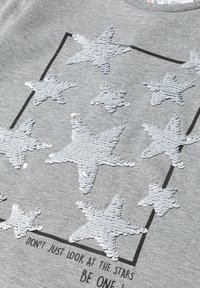 Grau meliertes T-Shirt aus Stoff mit silbernen Paillettensternen in verschiedenen Größen, umrandet von einem schwarzen rechteckigen Rahmen und schwarzem Text darunter.