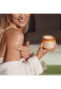 AHAVA Dead Sea Plants kroppsskrubb i en orange burk, visad med krämig konsistens applicerad på axeln. Modell iklädd en vit badrock.