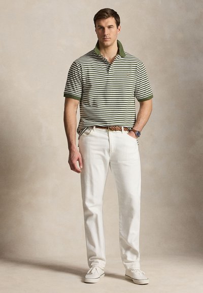 Άνδρας που φοράει πράσινη και λευκή ριγωτή πόλο, λευκό παντελόνι, καφέ πλεκτή ζώνη, λευκά αθλητικά παπούτσια και ρολόι χειρός, στέκεται μπροστά σε μπεζ φόντο.