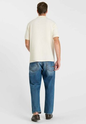 Man draagt een crèmekleurig shirt met korte mouwen, blauwe spijkerbroek en donkerbruine schoenen, staand met zijn rug naar de camera tegenover een effen witte achtergrond.