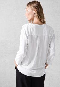 Blusa blanca de manga larga, tela suave, escote redondo y un sutil detalle de pliegue en la parte posterior, combinada con pantalones negros.