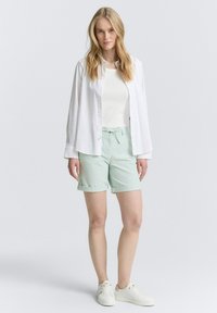 Shorts en coton vert clair avec des ourlets retroussés, associés à un débardeur blanc et une chemise blanche déboutonnée. Des baskets blanches complètent le look.
